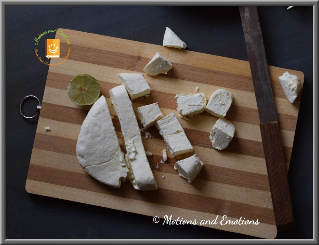 Paneer cubes cut from paneer block scattered on wooden board along with knife and lemon, top view