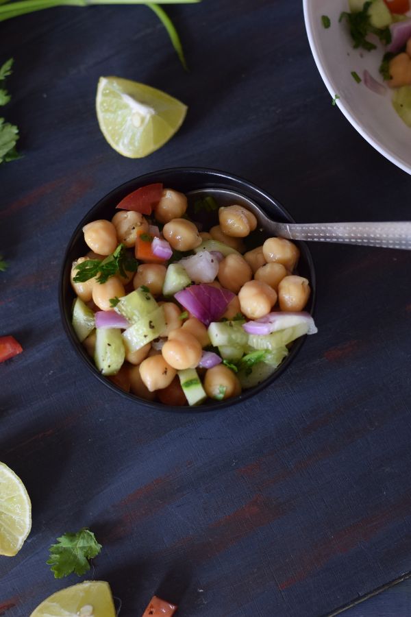 chickpea salad in a small black bowl with spoon and coriander leaves lemon in the background