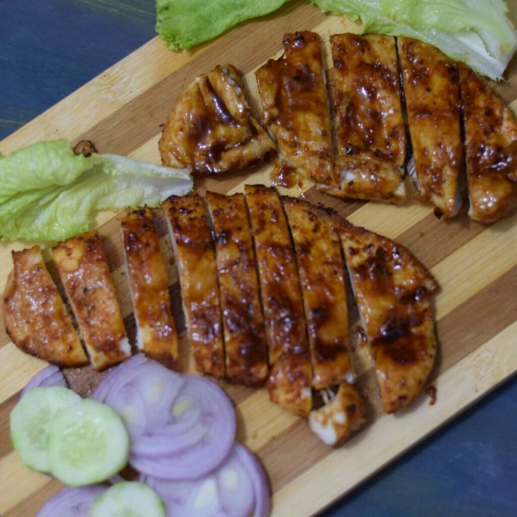 sliced air fried bbq chicken breasts on wooden board along with lettuce leaves and onion