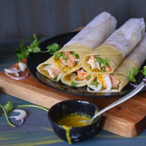 chicken kathi rolls stacked on black plate along with mustard sauce, onion and coriander leaves in the background