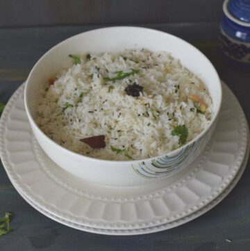 Jeera Rice in a white bowl kept on white plate along with cumin seeds and coriander leaves on the background