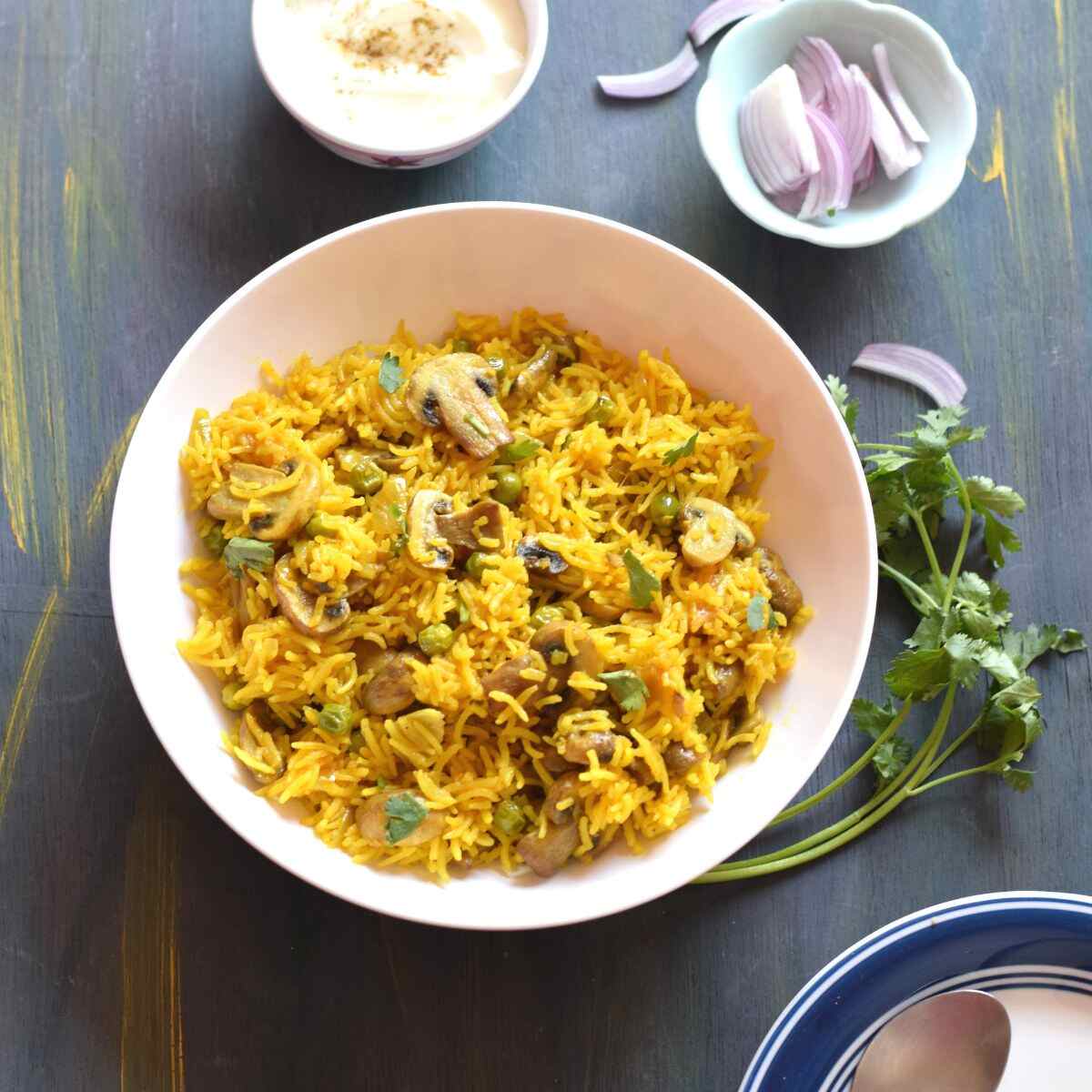 mushroom pilau rice in a bowl along with sliced onions, coriander leaves and a bowl of curd