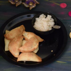 baked gujiyas in a black plate along with coconut stuffing in a bowl and colors and lamps in the background