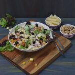 pasta salad in a large bowl kept on wooden board along with lettuce, grilled chicken and pasta