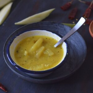 green mango dal in a white bowl kept on stoneware plate and mango strips and dry red chilli in the background