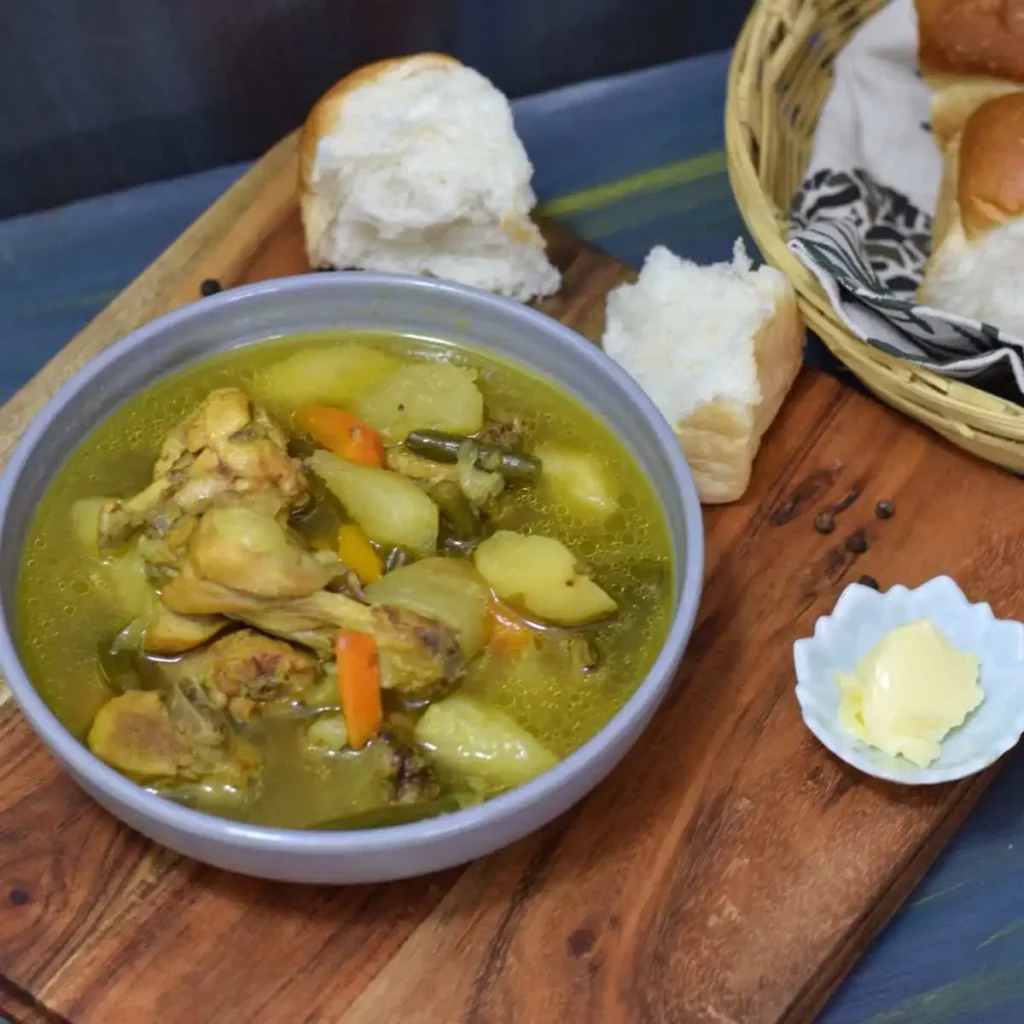 A bowl full of chicken stew kept on a wodden board along with butter and dinner rolls
