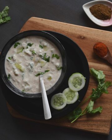 raita in a black bowl kept on a black plate & wooden board in the background