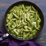 a pan full of green sauce pasta along with a blue cloth in the background