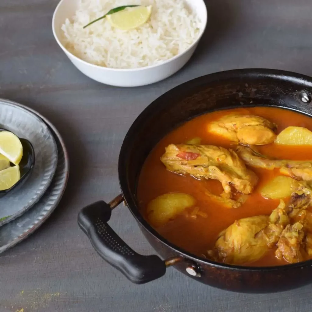 chicken curry in a wok, a bowl of rice and lemon wedges in the background