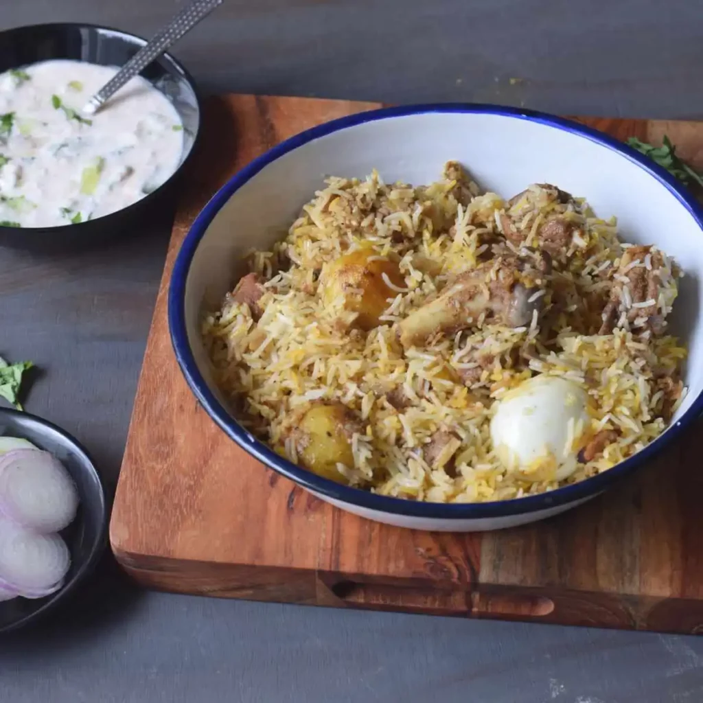 mutton biryani in a white dish kept on wooden board along with raita and sliced onion