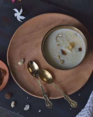Top view of a bowl of payesh kept in bronze bowl on wooden plate along with golden spoons