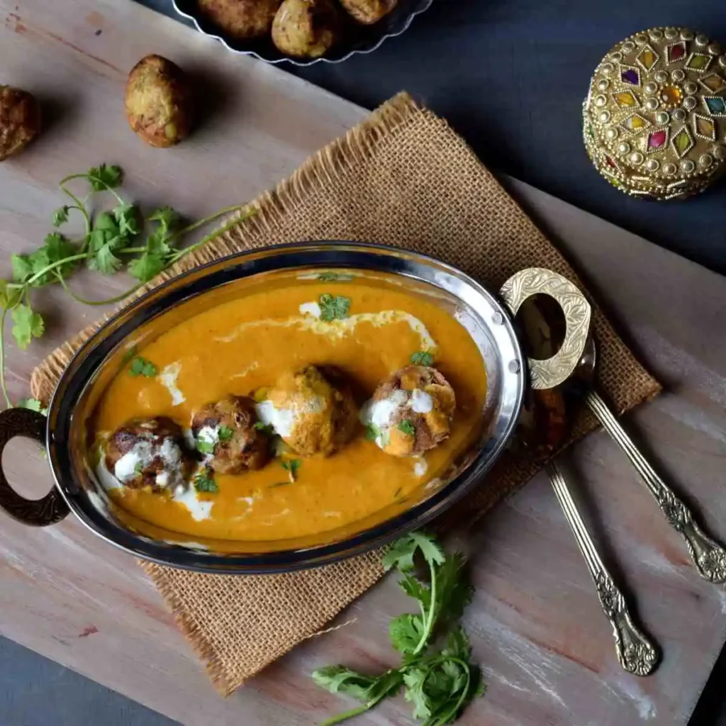 jackfruit kofta curry served in a steel server along with golden spoons, decorative items, koftas, coriander leaves in the background
