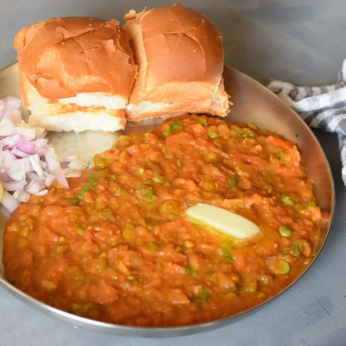 Pav Bhaji served on a steel plate with a dollop of butter , chopped onion and lemon wedge
