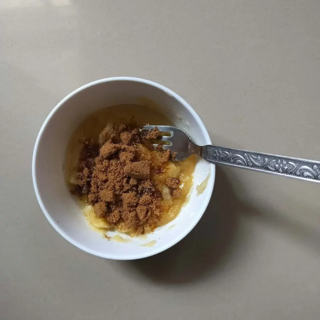 addition of brown sugar and oil into mashed banana in a bowl