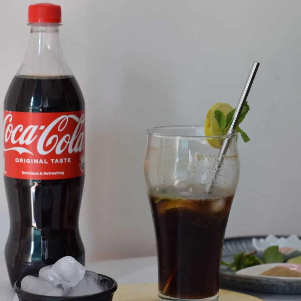 Masala coke in a glass with straw and coke bottle, ice cubes and spices in the background